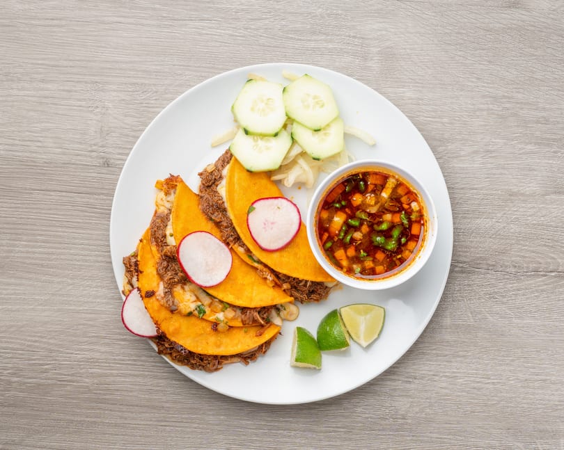 Birria tacos with radishes, cucumbers, onions, limes, and a side of consommé on a white plate.