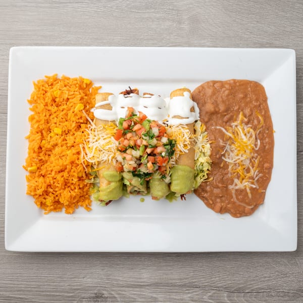 A white rectangular plate holds four taquitos topped with sour cream, pico de gallo, and guacamole, alongside orange rice and refried beans.