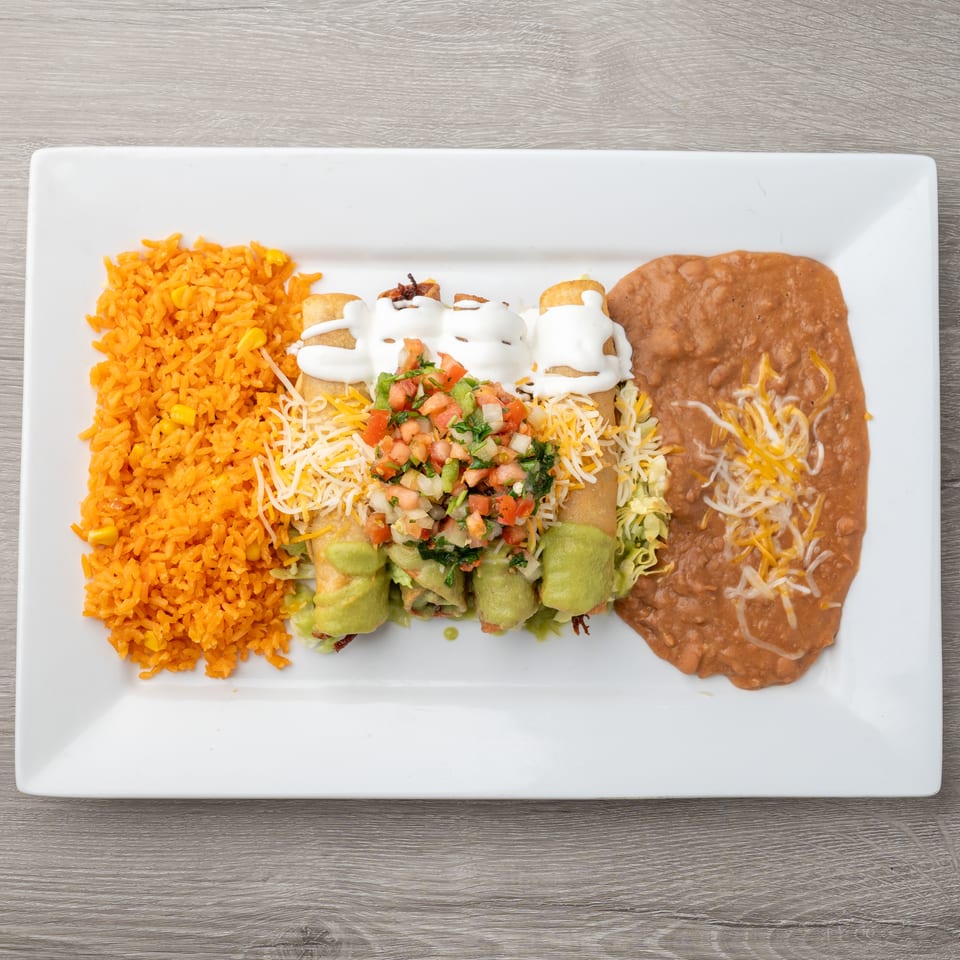 A white rectangular plate holds four taquitos topped with sour cream, pico de gallo, and guacamole, alongside orange rice and refried beans.