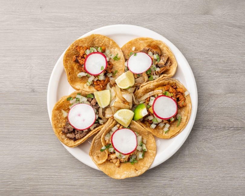 Five street tacos with various fillings, radishes, onions, cilantro, and lime wedges on a white plate.