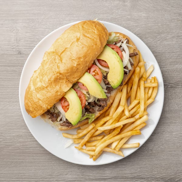 Steak sandwich with avocado, tomato, onion, and lettuce, served with french fries on a white plate.