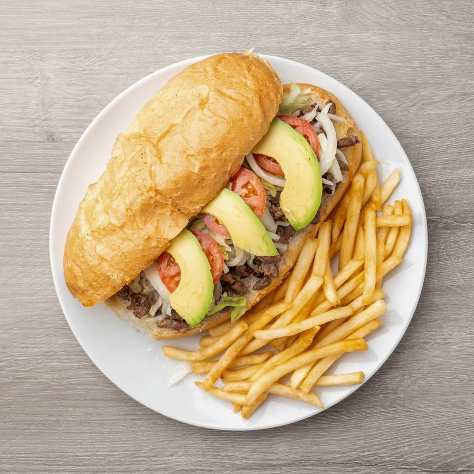 Steak sandwich with avocado, tomato, onion, and lettuce, served with french fries on a white plate.