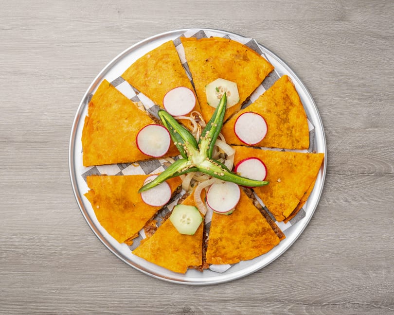 Golden quesadillas arranged in a circle on a metal tray, garnished with radishes, cucumbers, and jalapeños.