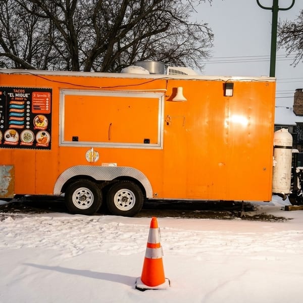 An orange food truck, "Tacos El Mibue," sits in a snowy parking lot with a white car nearby.