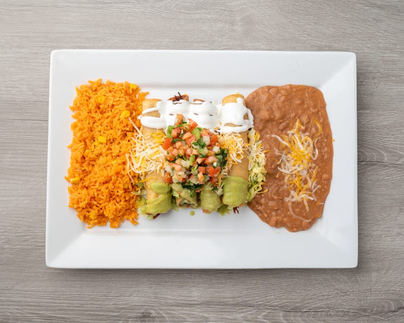 A white rectangular plate holds four taquitos with sour cream, pico de gallo, rice, and refried beans.