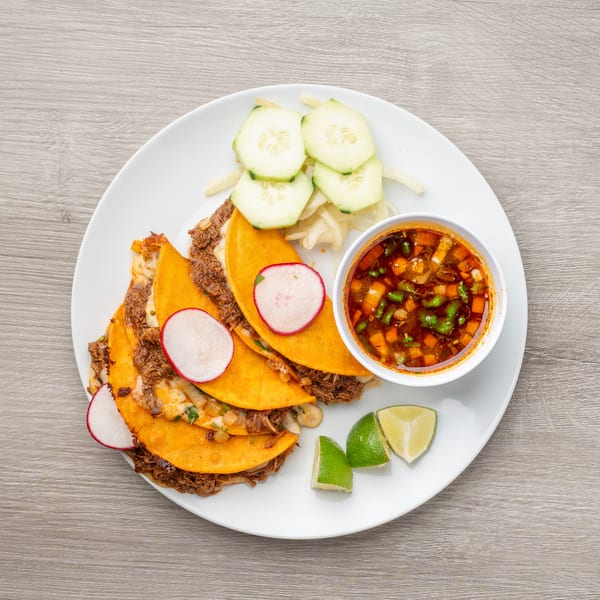 Birria tacos with radishes, cucumbers, limes, and a side of consommé on a white plate.
