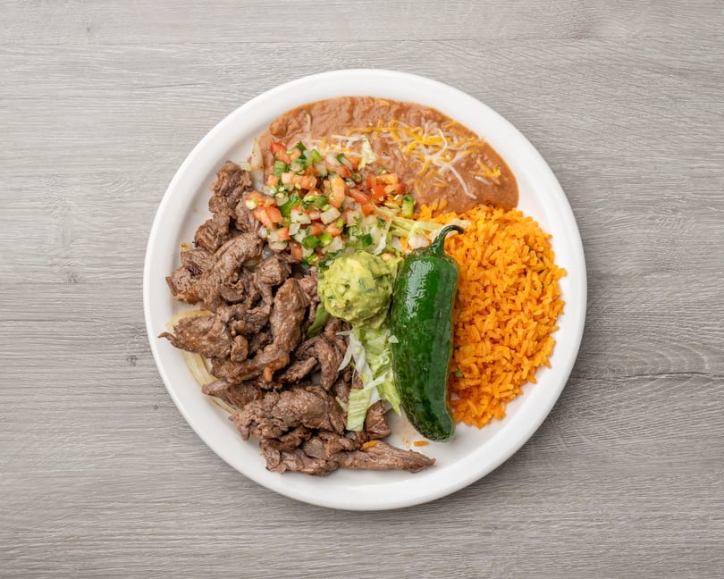 A Mexican dish with sliced steak, rice, refried beans, guacamole, pico de gallo, and a whole jalapeño.