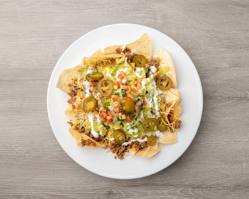 A white plate filled with nachos topped with ground beef, cheese, lettuce, salsa, sour cream, and jalapeños.