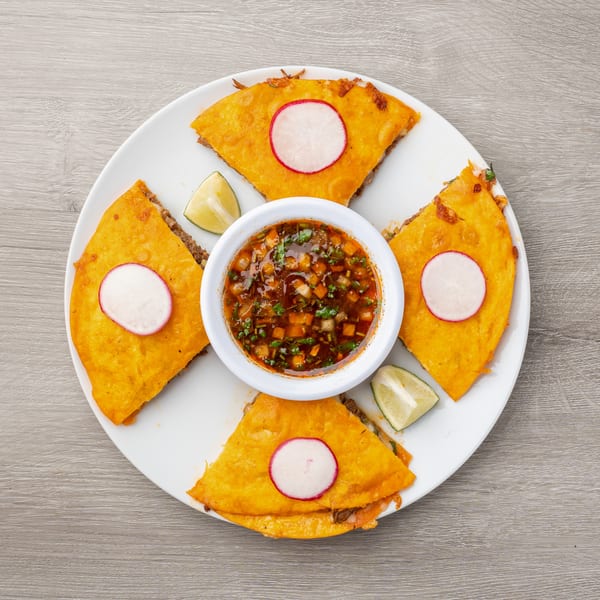 Four golden-brown quesadillas with radish slices and lime wedges surround a bowl of dipping sauce.